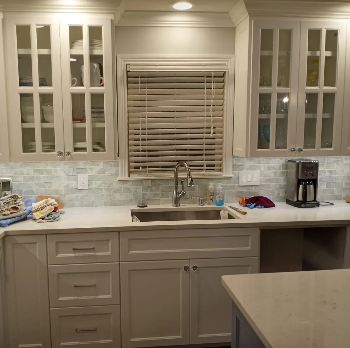 Finished white custom kitchen cabinetry with glass-front upper cabinets, sink wall, tile backsplash, and fitted lower storage